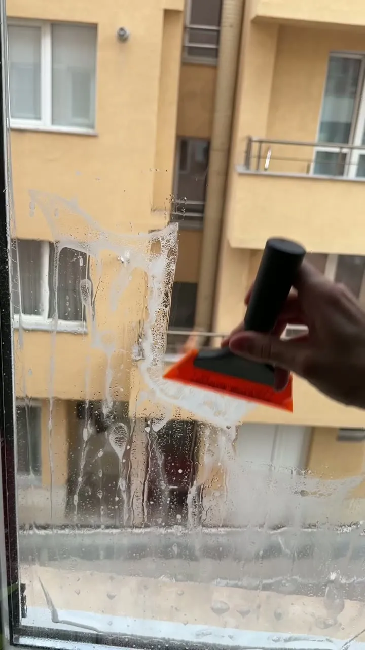 Ръка, прекарваща ракел по мокро фолио на прозорец, с двор на жилищна сграда зад стъклото